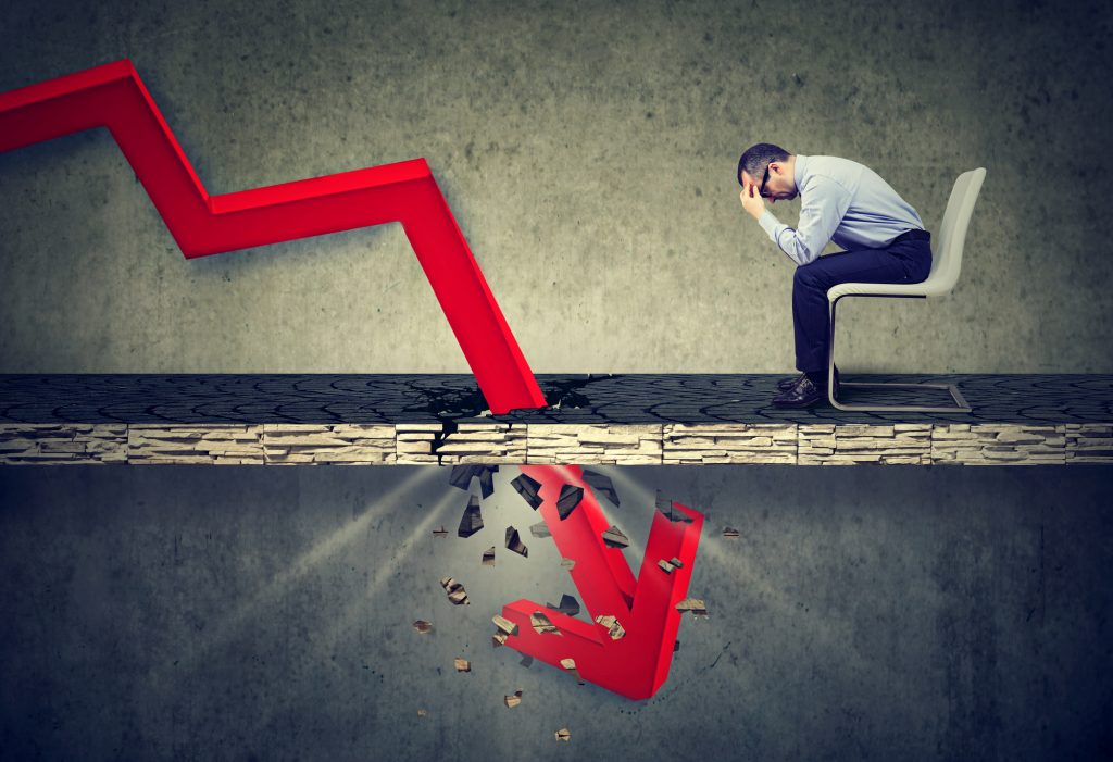 A man sitting in a chair, holding his forehead, looking stressed with a downward arrow signifying bankruptcy.