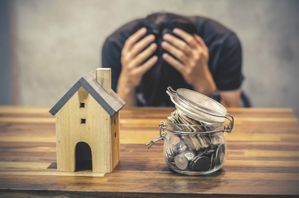 A man holding his head in distress with a miniature house and coins in a small jar, symbolizing financial issues.