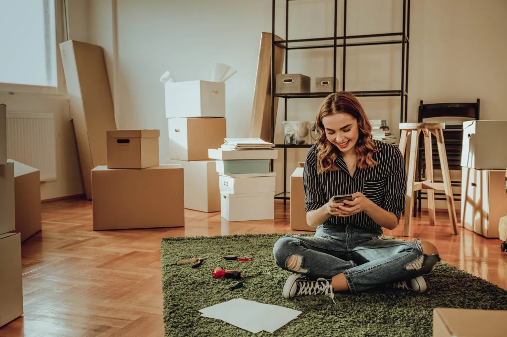 A woman sitting on the floor surrounded by moving boxes, smiling while using her cellphone, symbolizing a smooth relocation process.